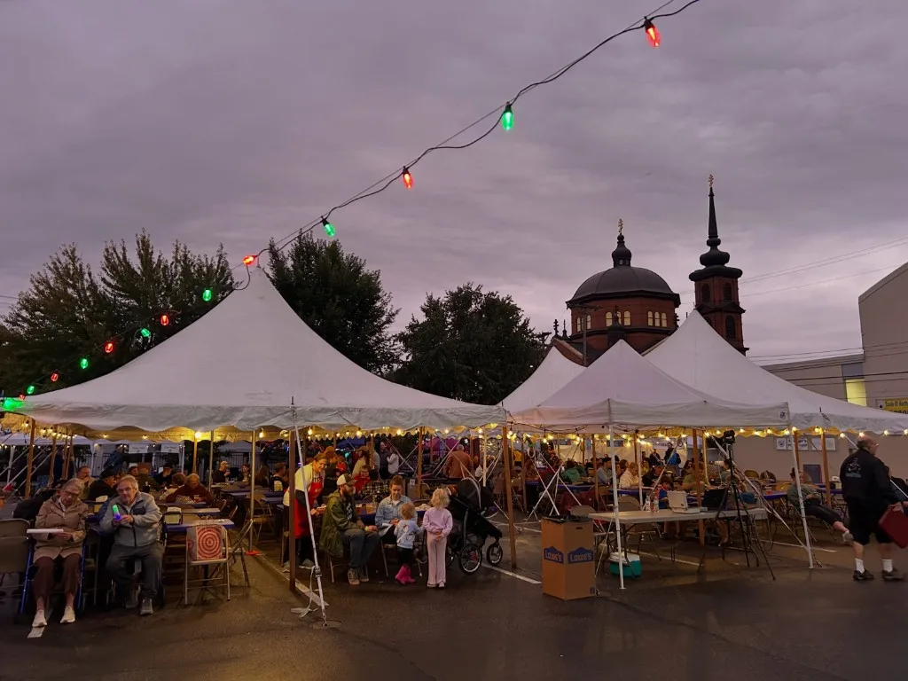 White festival tents and colored string lights at dusk, with the cathedral towers behind a full lot