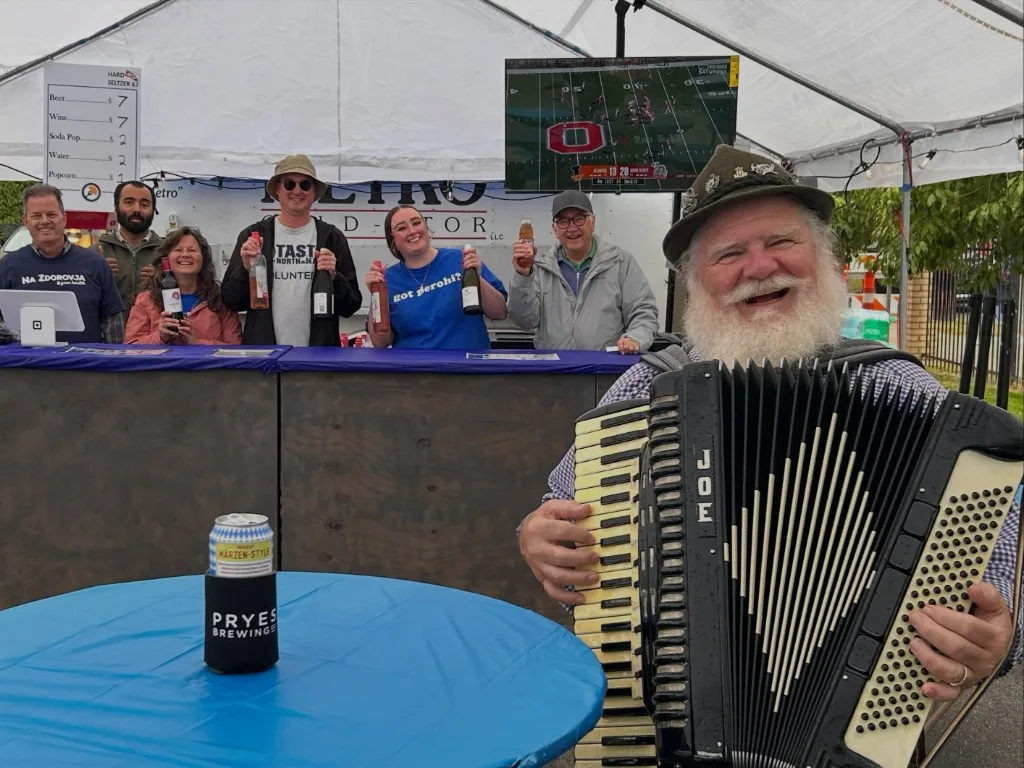 Joe Macko smiling with his accordion in front of the drink tent, volunteers behind the bar