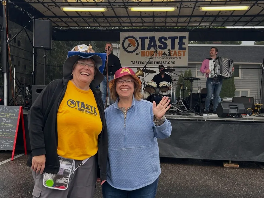 Two guests smiling in front of the outdoor stage, one in a festival shirt, with the band and the Taste of Northeast banner behind them